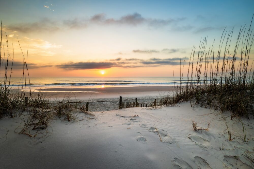 Sunrise over the Atlantic Ocean at Amelia Island, Florida, with sea oats and a weathered dune fence framing a sandy beach and the sun reflecting in the wet sand.