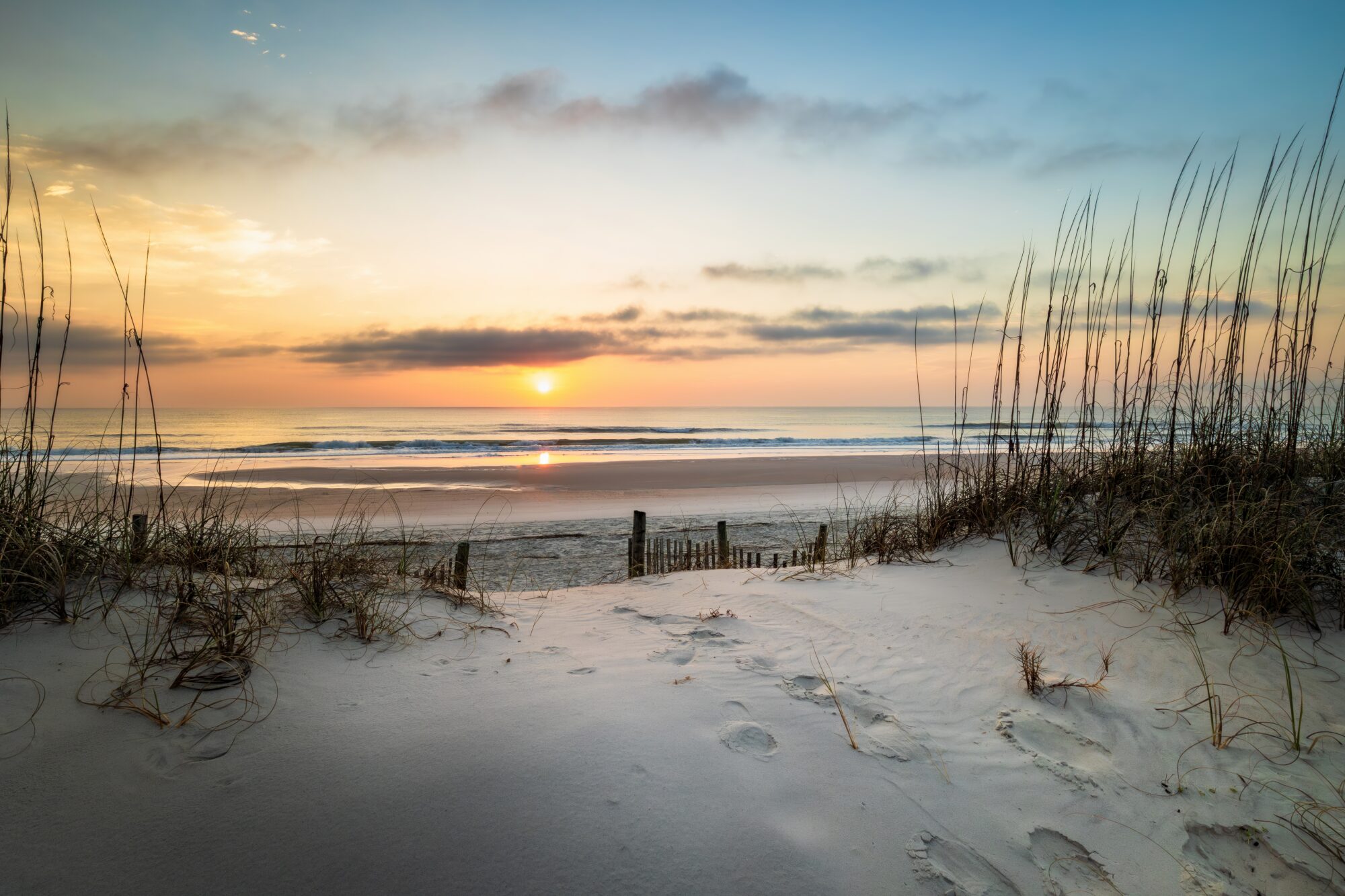 Sunrise over the Atlantic Ocean at Amelia Island, Florida, with sea oats and a weathered dune fence framing a sandy beach and the sun reflecting in the wet sand.