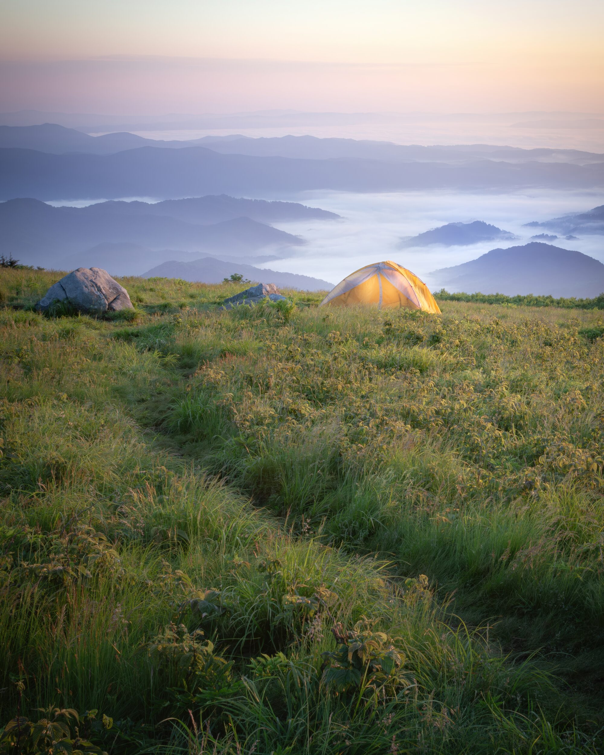 A glowing tent on an open grassy bald at sunrise in the Roan Highlands, with layered blue ridges fading into the distance.